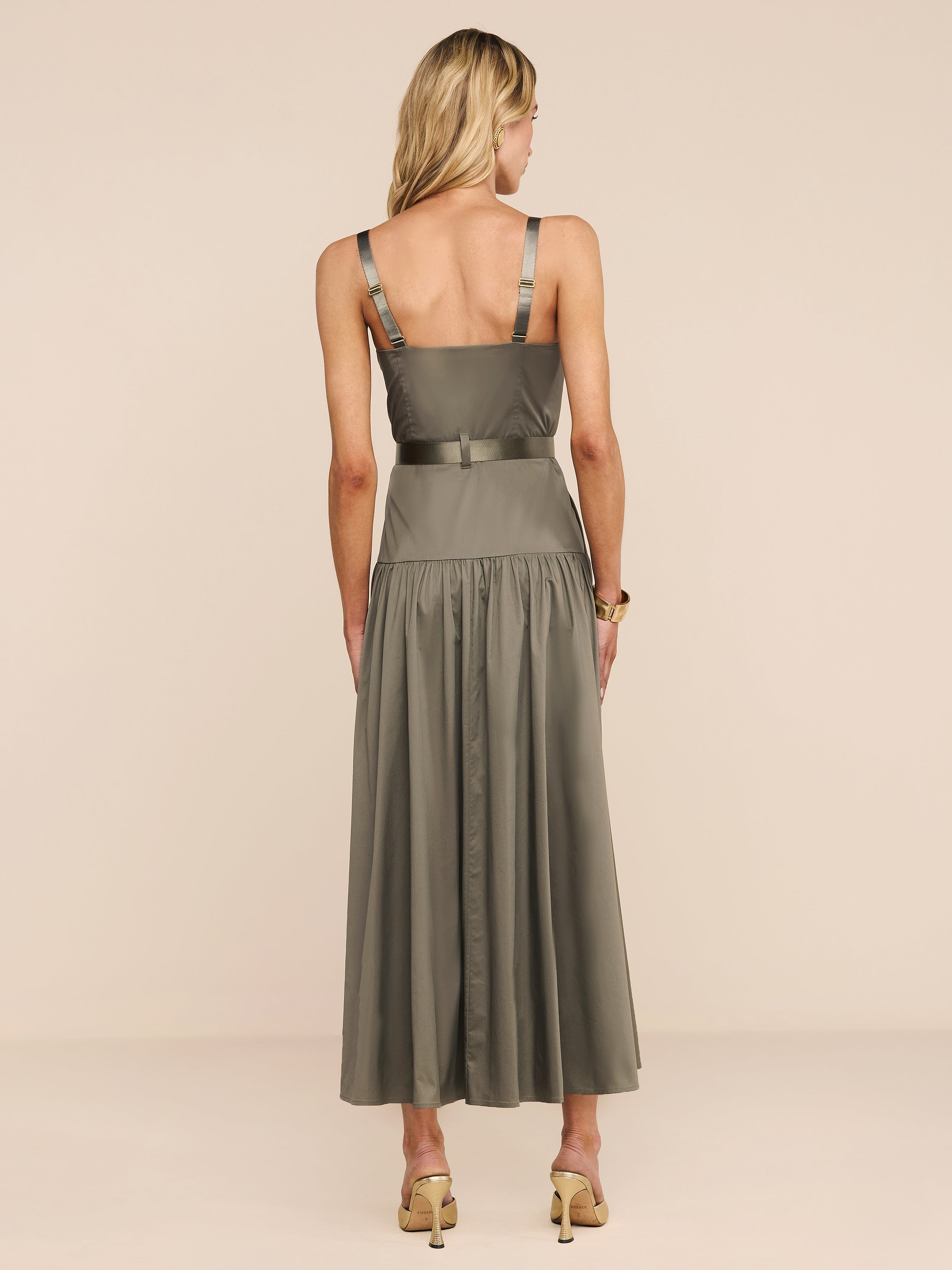 A woman with blonde hair faces away, wearing the Rima Dress—an olive green maxi with a fitted bodice, flared skirt, and adjustable straps—paired with beige high-heeled sandals against a plain beige background.