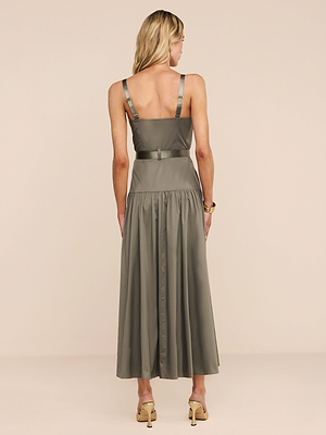 A woman with blonde hair faces away, wearing the Rima Dress—an olive green maxi with a fitted bodice, flared skirt, and adjustable straps—paired with beige high-heeled sandals against a plain beige background.
