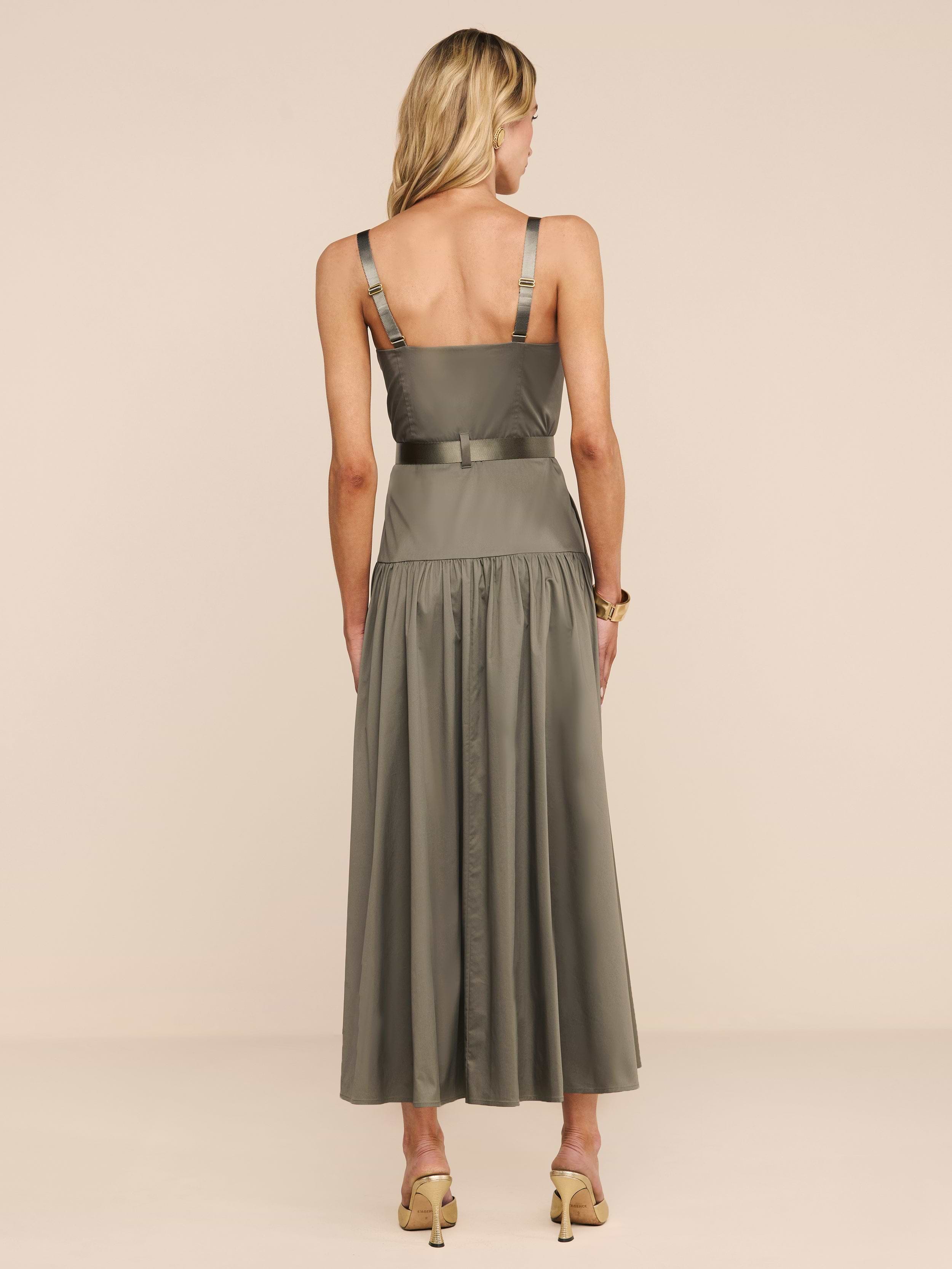 A woman with blonde hair faces away, wearing the Rima Dress—an olive green maxi with a fitted bodice, flared skirt, and adjustable straps—paired with beige high-heeled sandals against a plain beige background.