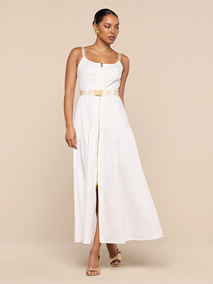 A woman models the Rima Dress, a sleeveless, ankle-length white maxi made from stretch poplin with a front zipper and cream belt. She pairs it with gold earrings and heeled sandals, set against a beige background.