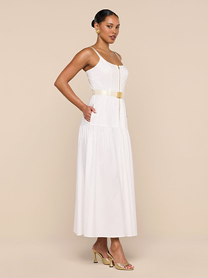 A woman stands sideways against a plain beige background, wearing the Rima Dress—a sleeveless white stretch poplin maxi—styled with a gold belt, gold high-heeled sandals, and gold earrings.