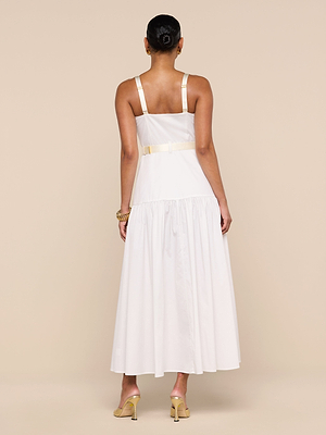 A woman faces away, wearing the Rima Dress—a sleeveless white stretch poplin maxi with a fitted bodice, wide straps, gathered skirt, and light belt. Her hair is up in a bun, styled with gold heels and a bracelet.