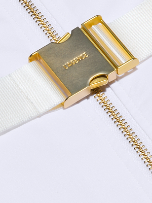 Close-up of a shiny gold metal HERMÈS buckle on a white strap, part of the Rima Dress, resting on a white surface with a vertical gold zipper underneath.