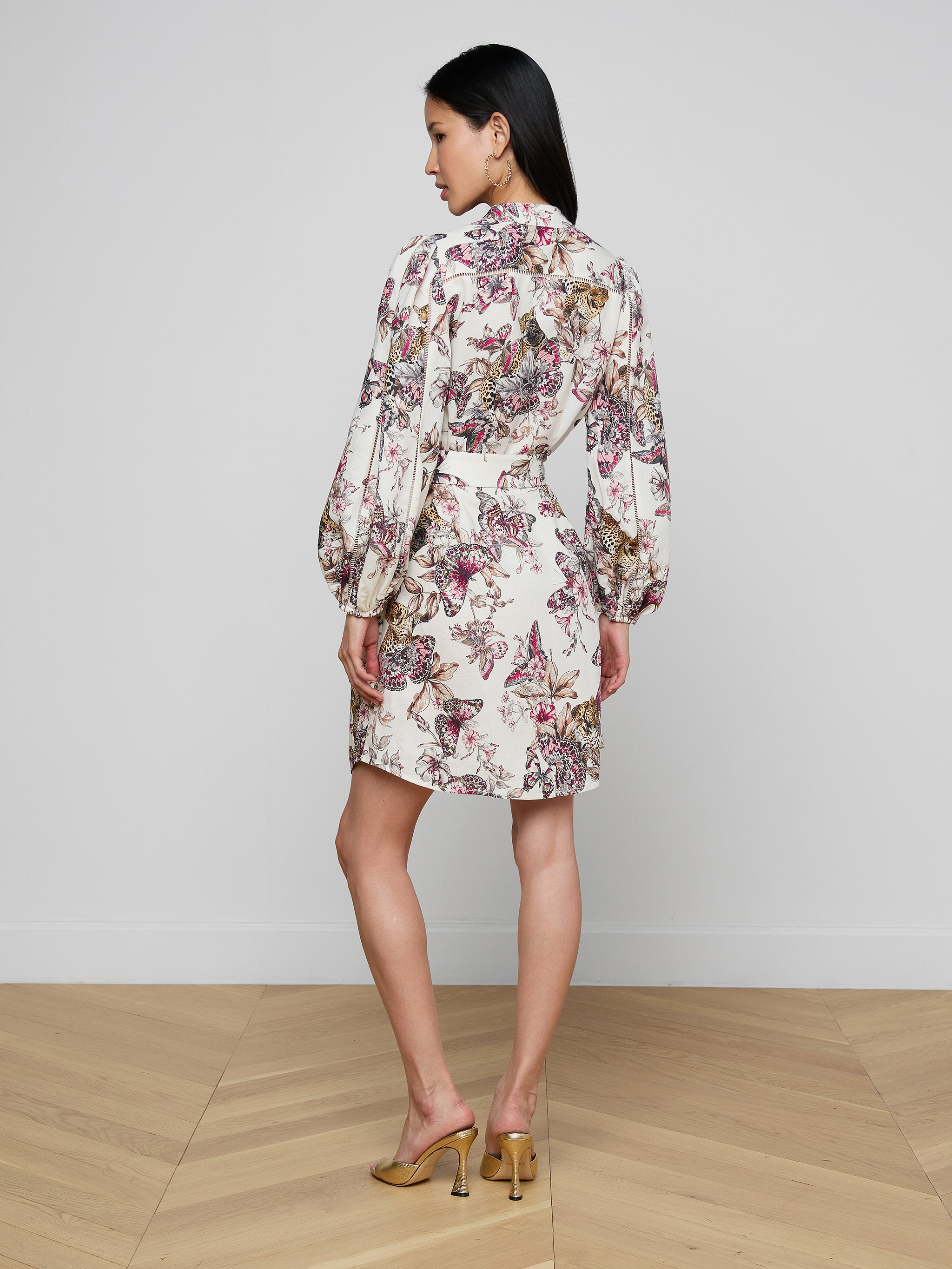 A woman stands facing away against a plain white background, wearing the Kiersten Dress—a white puffed sleeve dress with a floral pattern—and tan high heels.