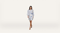 A woman wearing a light blue and white floral dress with long sleeves walks forward against a plain white background. She is wearing open-toed heels and has loose, wavy hair.