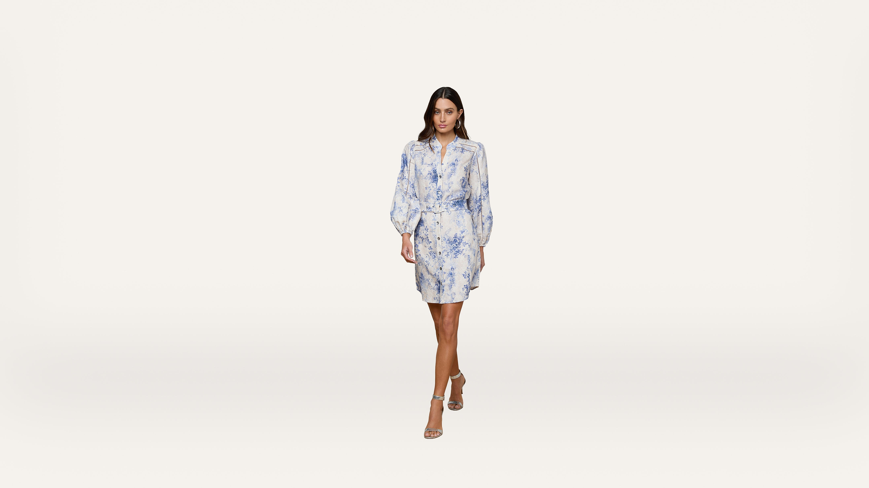 A woman wearing a light blue and white floral dress with long sleeves walks forward against a plain white background. She is wearing open-toed heels and has loose, wavy hair.