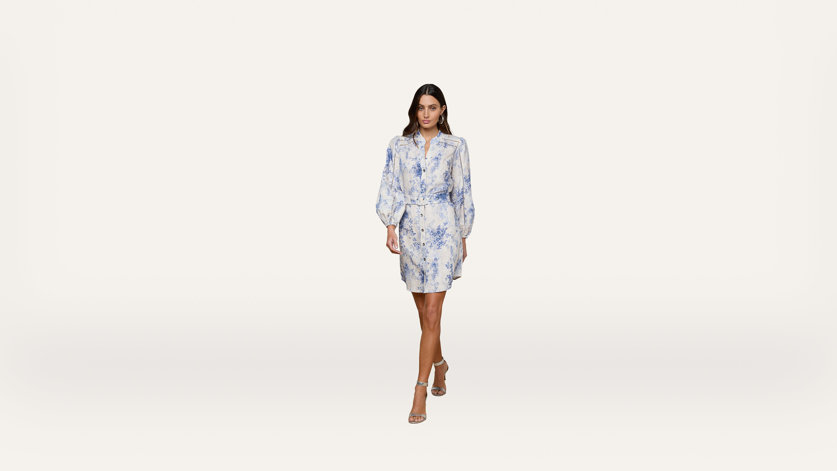 A woman wearing a light blue and white floral dress with long sleeves walks forward against a plain white background. She is wearing open-toed heels and has loose, wavy hair.