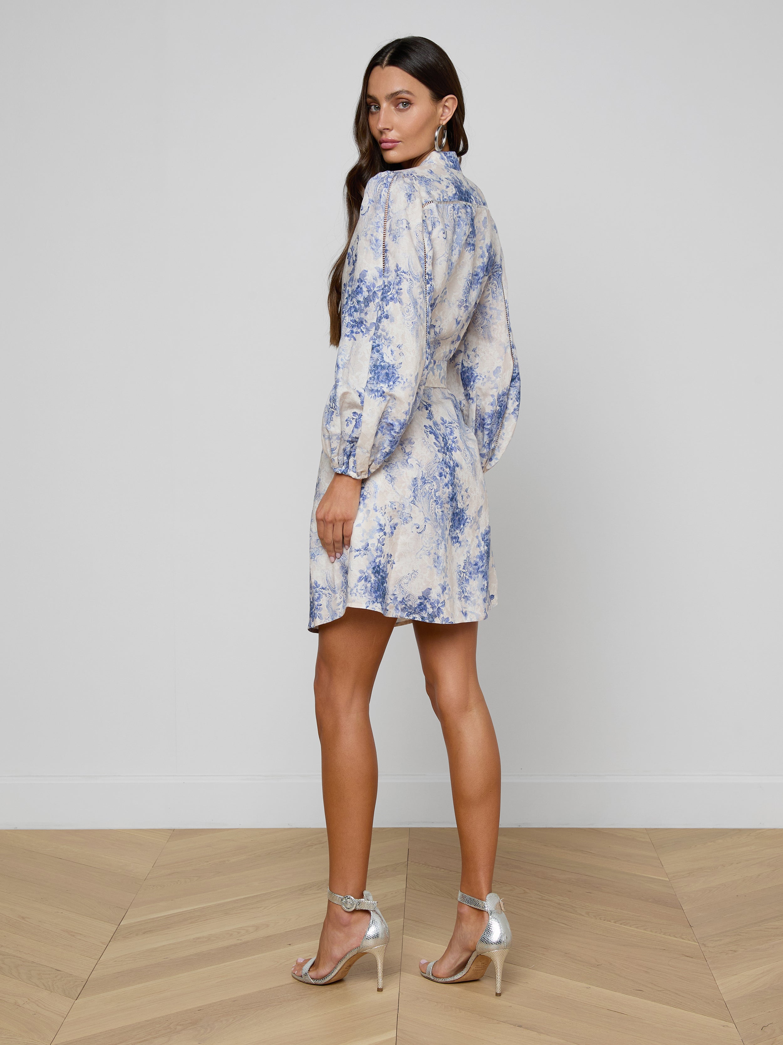 A woman with long brown hair stands on a wooden floor, facing slightly away from the camera against a white wall, wearing the Kiersten Dress—a standout print linen design with blouson sleeves—and silver high-heeled sandals.