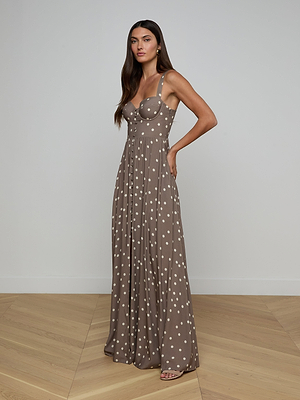 A woman poses confidently indoors, wearing the Rosette Maxi Dress—a taupe, white polka dot summer dress with a fitted bodice, wide straps, and a flowing maxi skirt. She has long straight hair and stands with one hand on her hip.