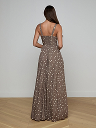 A woman with long brown hair faces away, wearing the Rosette Maxi Dress—a sleeveless brown maxi dress with white polka dots, straps, and a fitted bodice. She stands on light wooden flooring against a plain light wall. Perfect for warm weather.