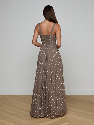 A woman with long brown hair faces away, wearing the Rosette Maxi Dress—a sleeveless brown maxi dress with white polka dots, straps, and a fitted bodice. She stands on light wooden flooring against a plain light wall. Perfect for warm weather.
