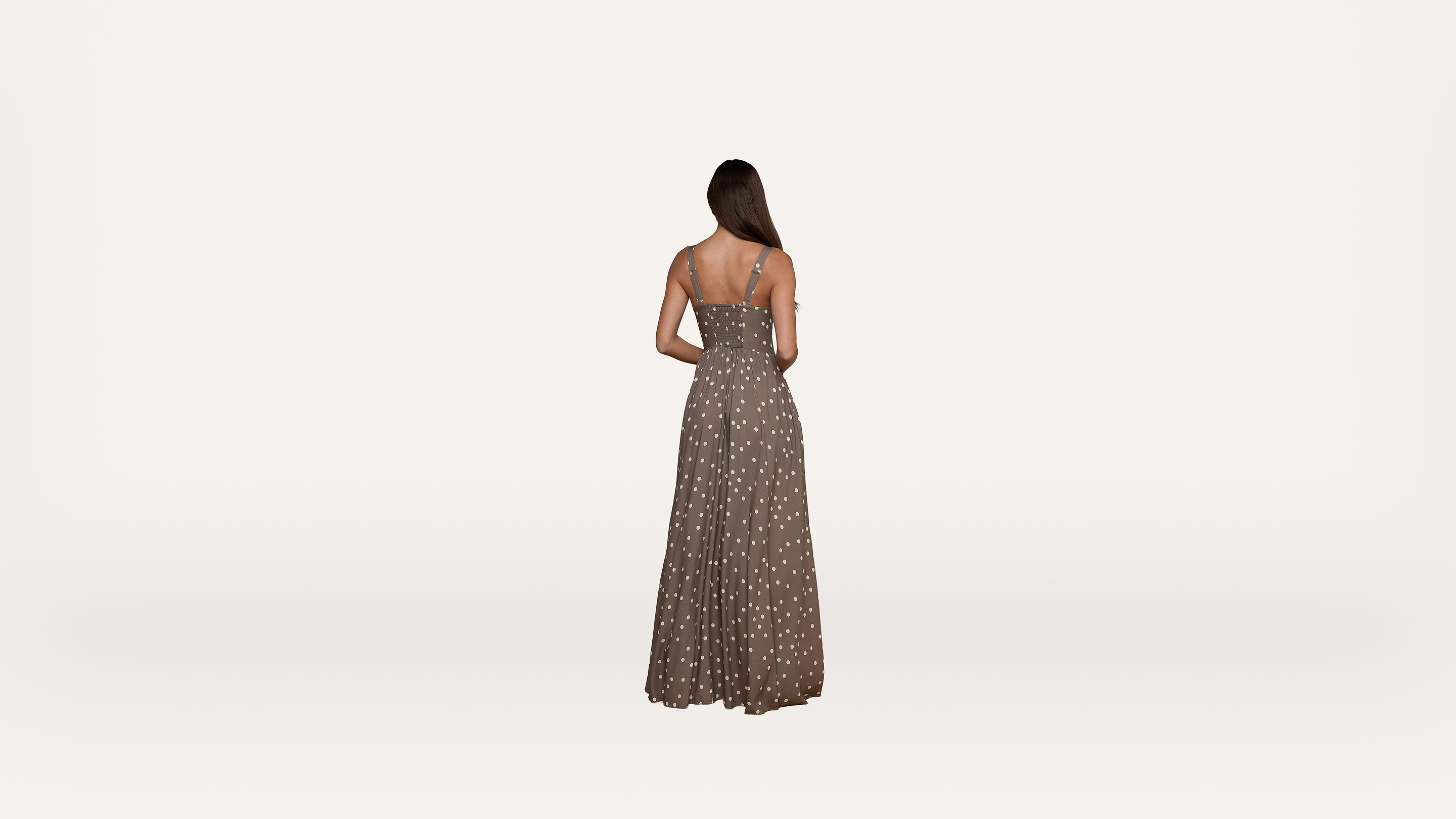 A woman with long hair, seen from behind, wears a sleeveless, brown maxi dress with white polka dots against a plain, light background.
