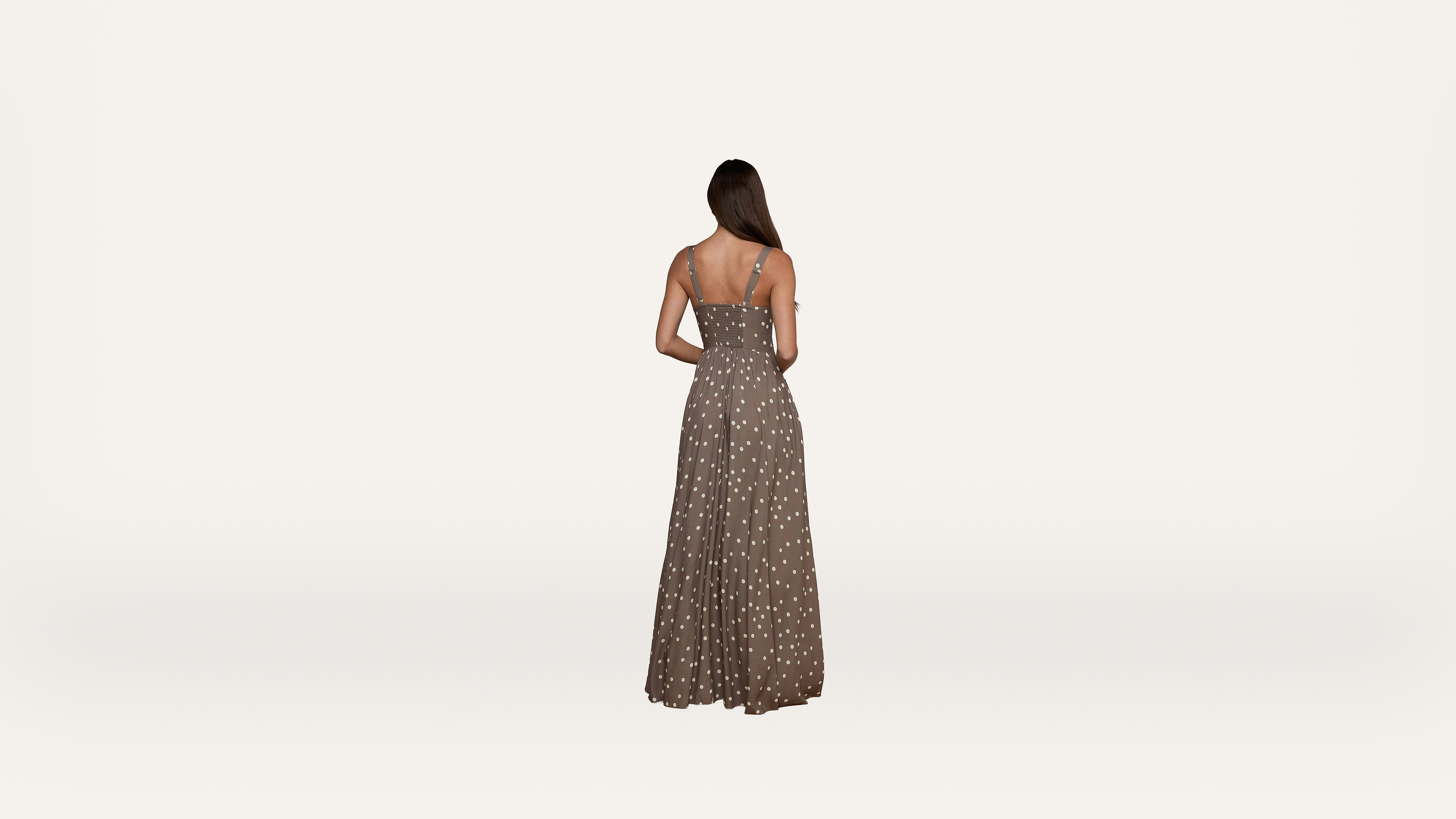 A woman with long hair, seen from behind, wears a sleeveless, brown maxi dress with white polka dots against a plain, light background.