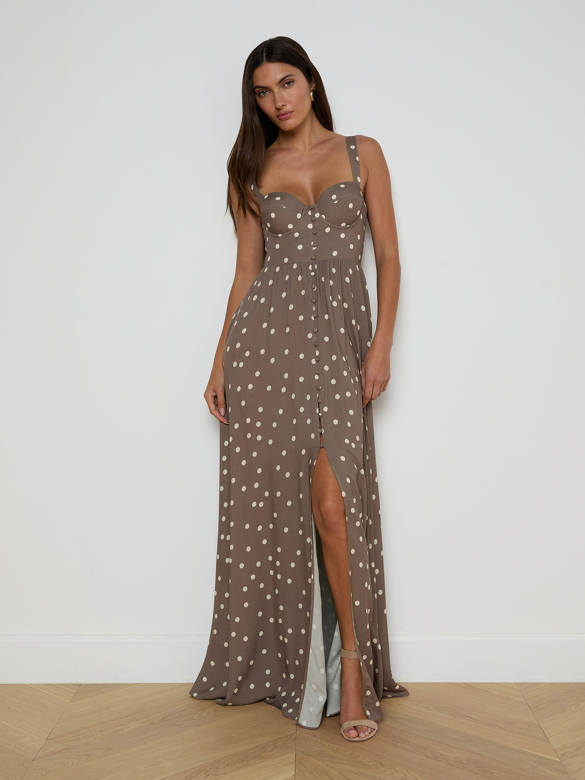 A woman with long dark hair poses by a white wall wearing the Rosette Maxi Dress—a taupe polka dot maxi with a fitted bodice and thigh-high slit—styled with beige high-heeled sandals for a chic, warm-weather look.