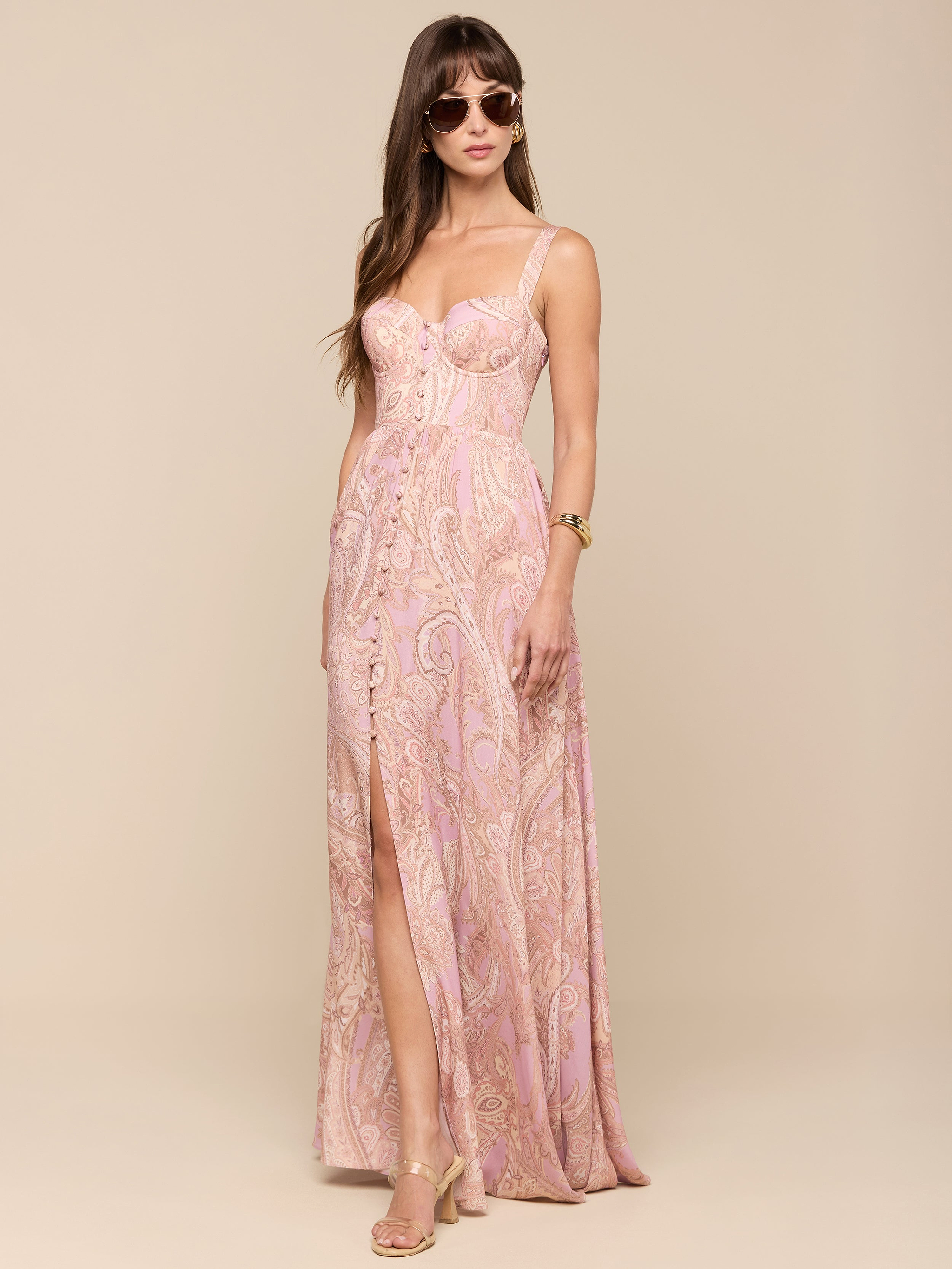 A woman stands against a plain white background wearing the Rosette Maxi Dress—a mauve paisley, sleeveless maxi dress with a thigh-high slit—paired with sunglasses, gold heels, and a watch.