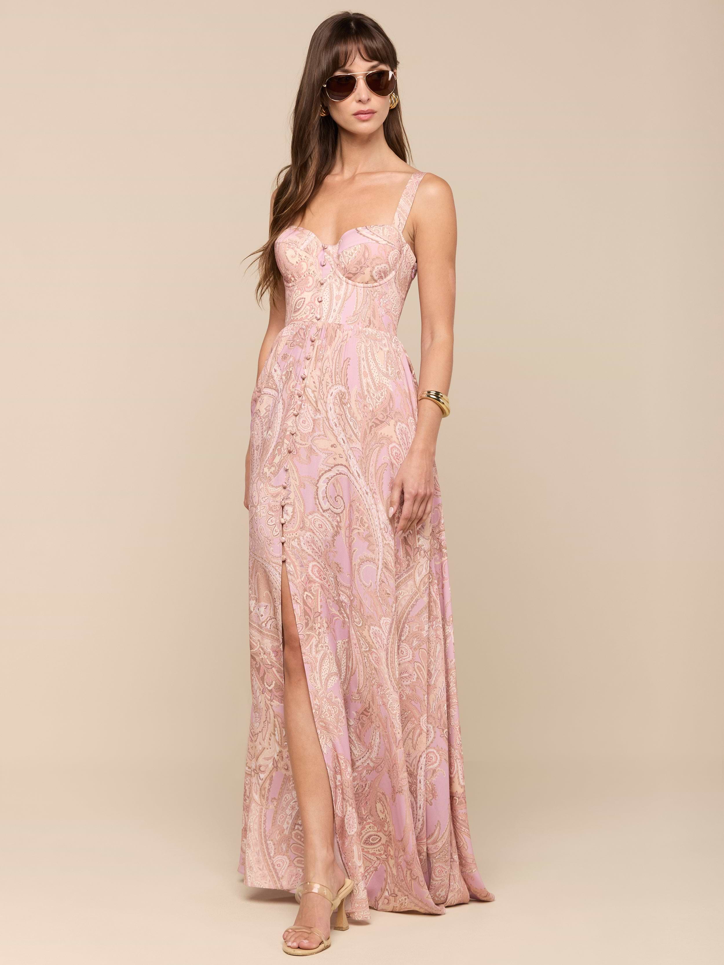 A woman stands against a plain white background wearing the Rosette Maxi Dress—a mauve paisley, sleeveless maxi dress with a thigh-high slit—paired with sunglasses, gold heels, and a watch.