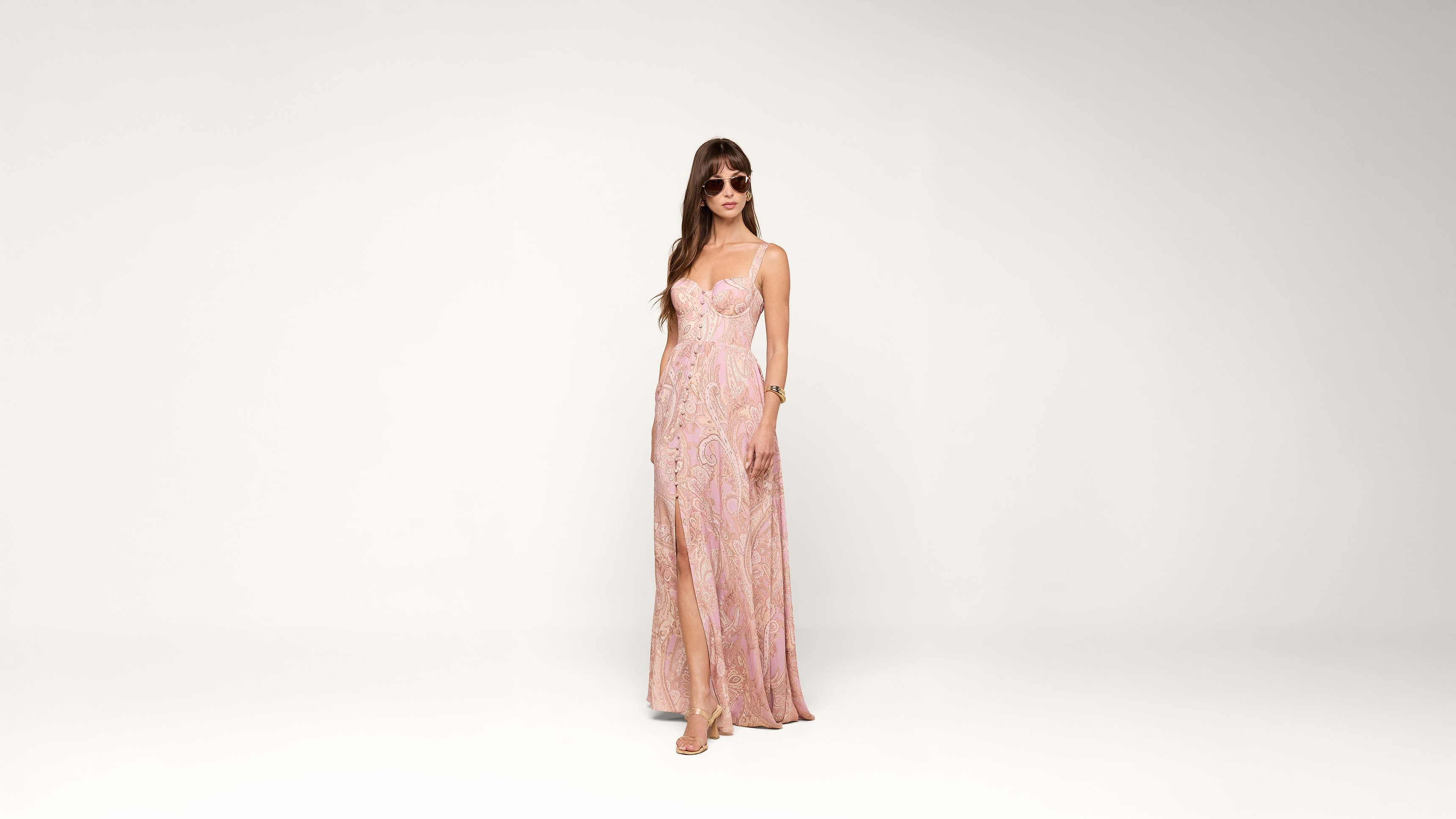 Woman wearing sunglasses and a light pink, floor-length sleeveless dress with a slit, standing against a plain white background.