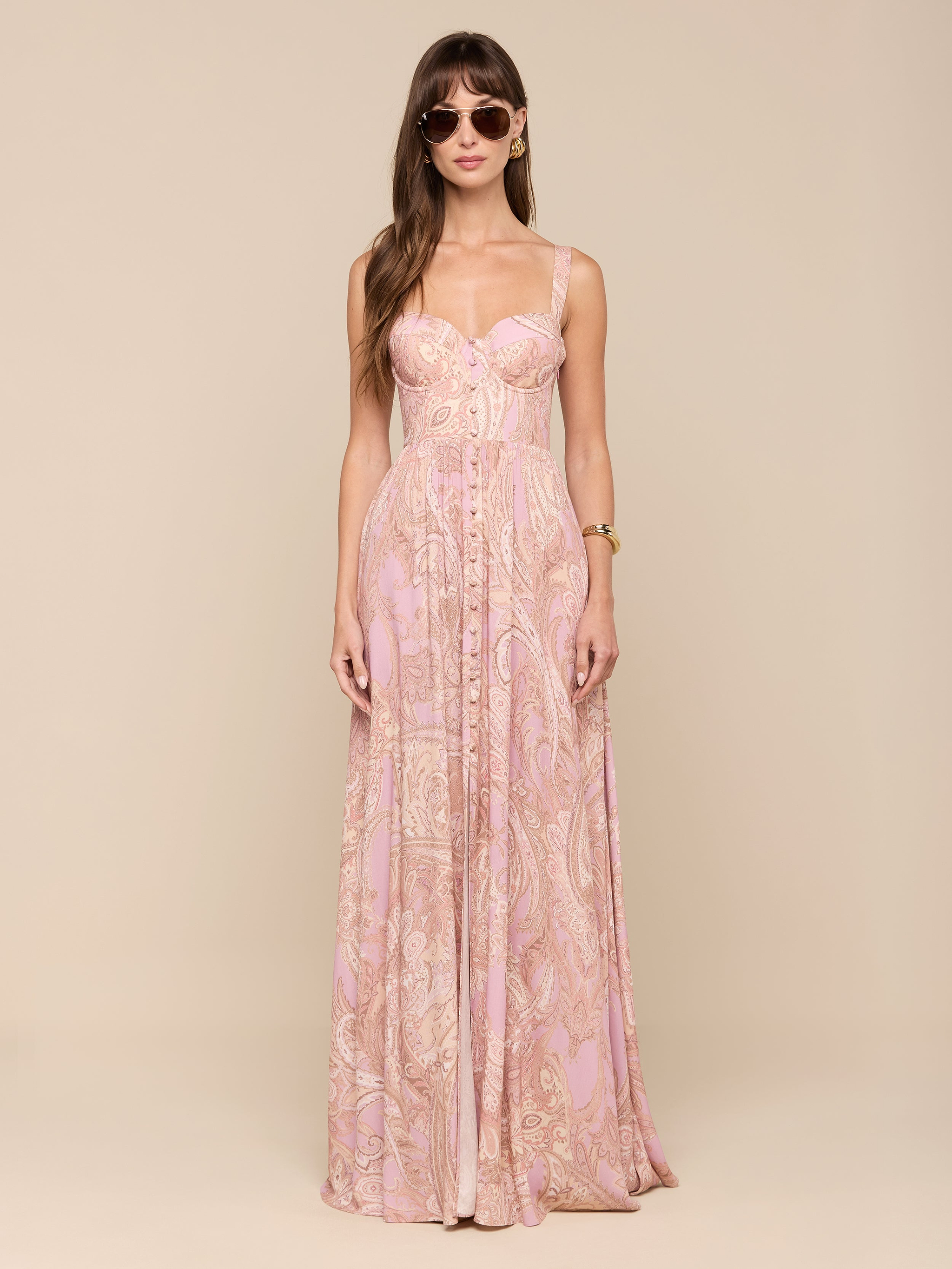 A woman with long brown hair stands facing forward against a plain white background, wearing sunglasses and the Rosette Maxi Dress—a long, mauve paisley maxi dress. Her arms are relaxed at her sides.