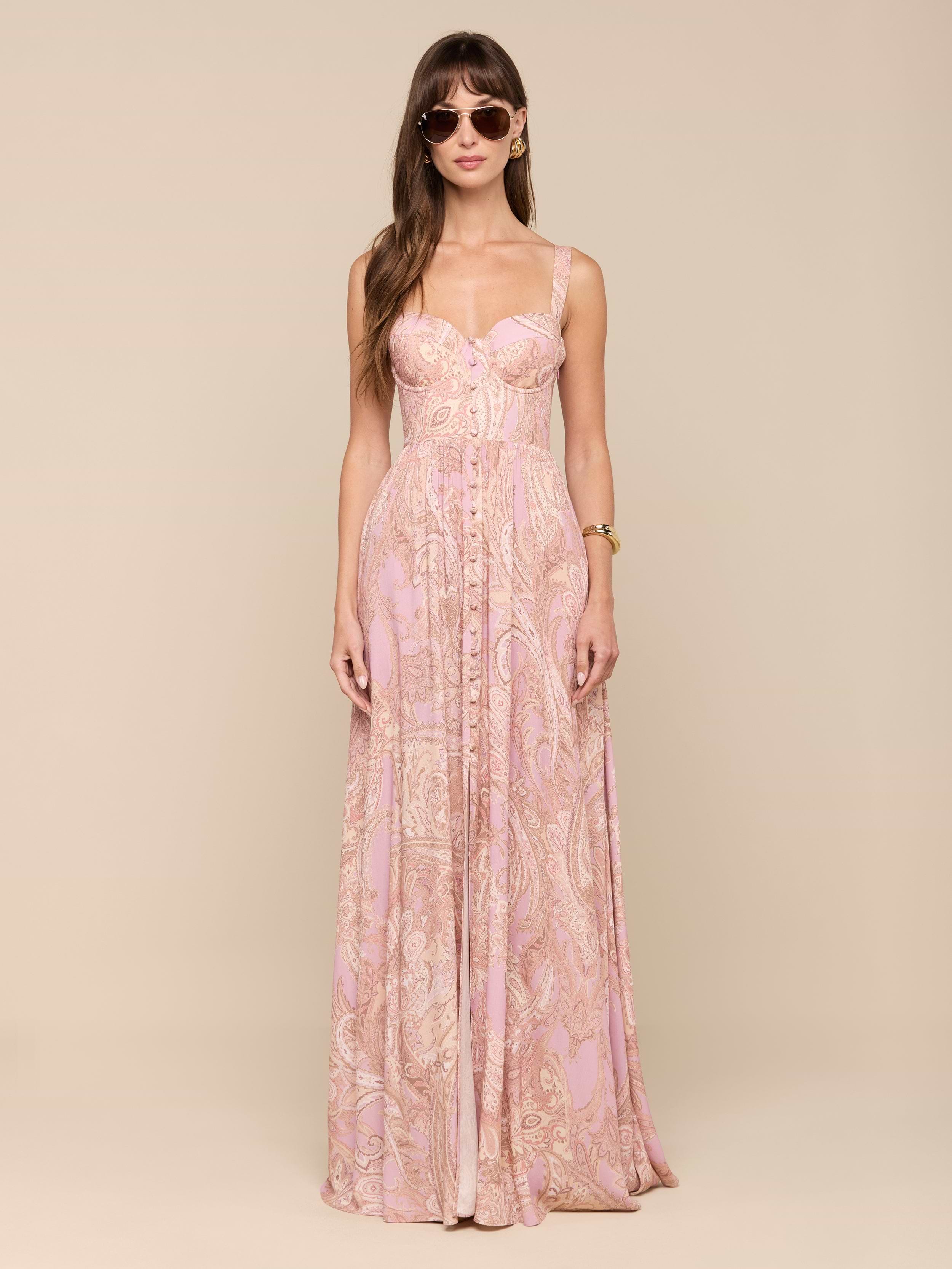 A woman with long brown hair stands facing forward against a plain white background, wearing sunglasses and the Rosette Maxi Dress—a long, mauve paisley maxi dress. Her arms are relaxed at her sides.