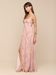 A woman with long brown hair stands in a relaxed pose against a plain white background, wearing the Rosette Maxi Dress—a light pink mauve paisley maxi dress featuring a slit. She accessorizes with a bracelet and gazes slightly downward.
