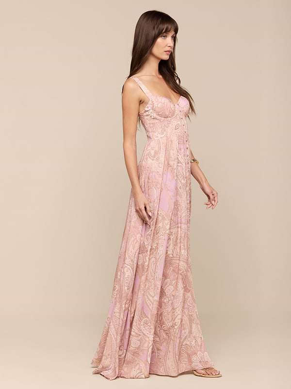 A woman in the Rosette Maxi Dress—a light pink, sleeveless maxi dress with a subtle mauve paisley pattern—stands against a plain white background, facing left.
