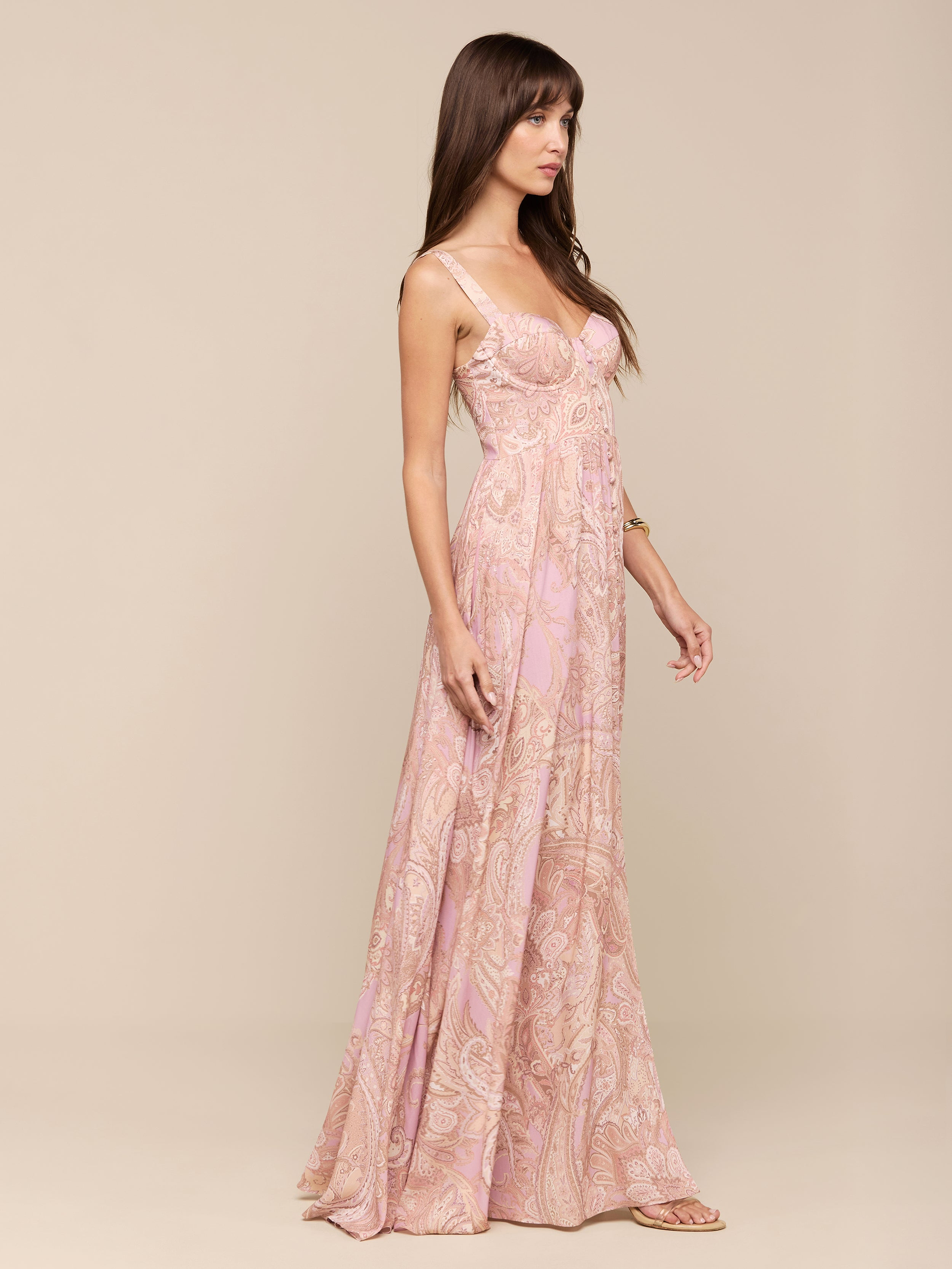 A woman in the Rosette Maxi Dress—a light pink, sleeveless maxi dress with a subtle mauve paisley pattern—stands against a plain white background, facing left.
