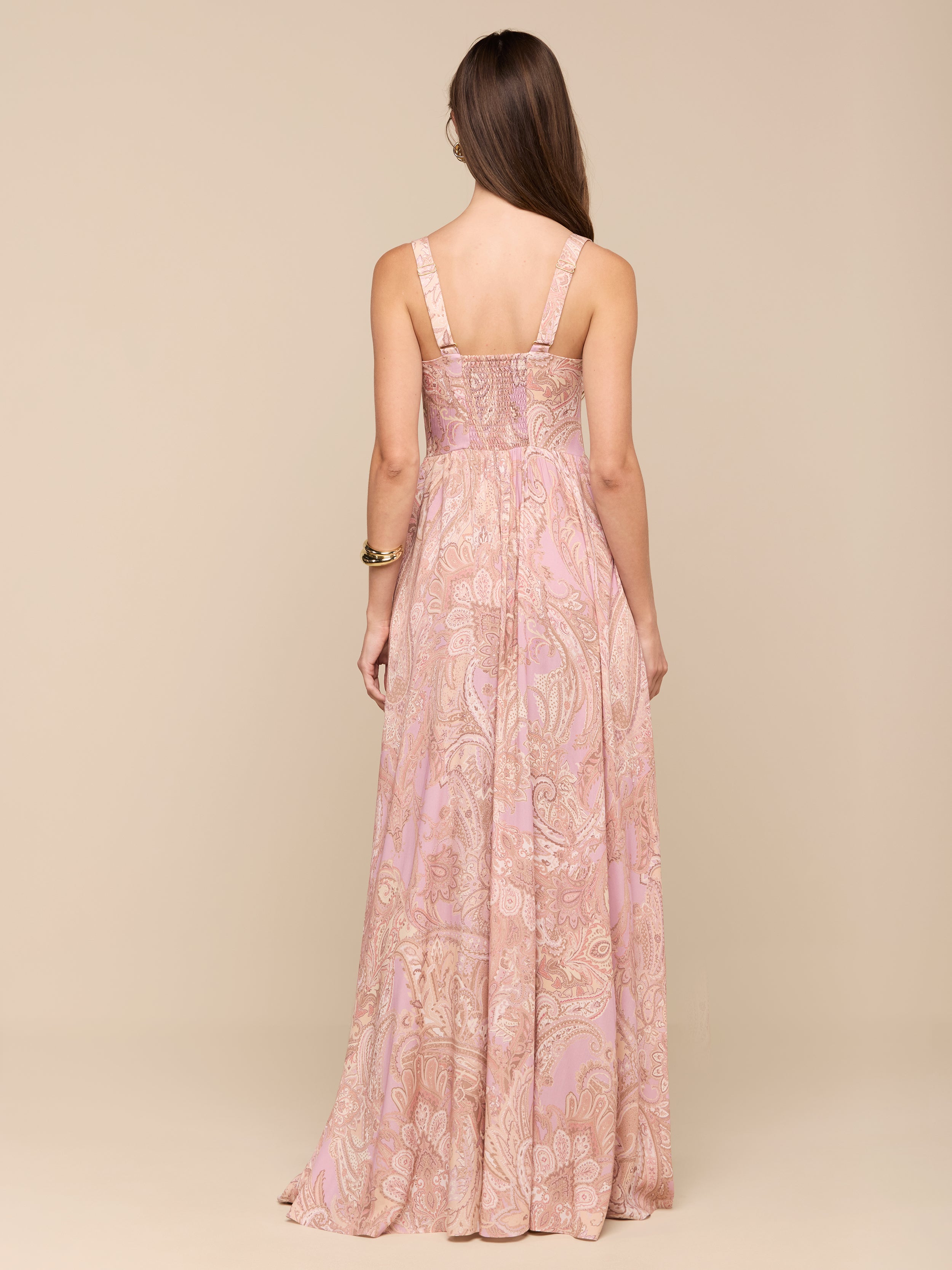 A woman with long brown hair stands with her back to the camera, wearing the Rosette Maxi Dress—a sleeveless, long mauve paisley maxi dress. She accessorizes with a bracelet on her right wrist, and the background is plain white.