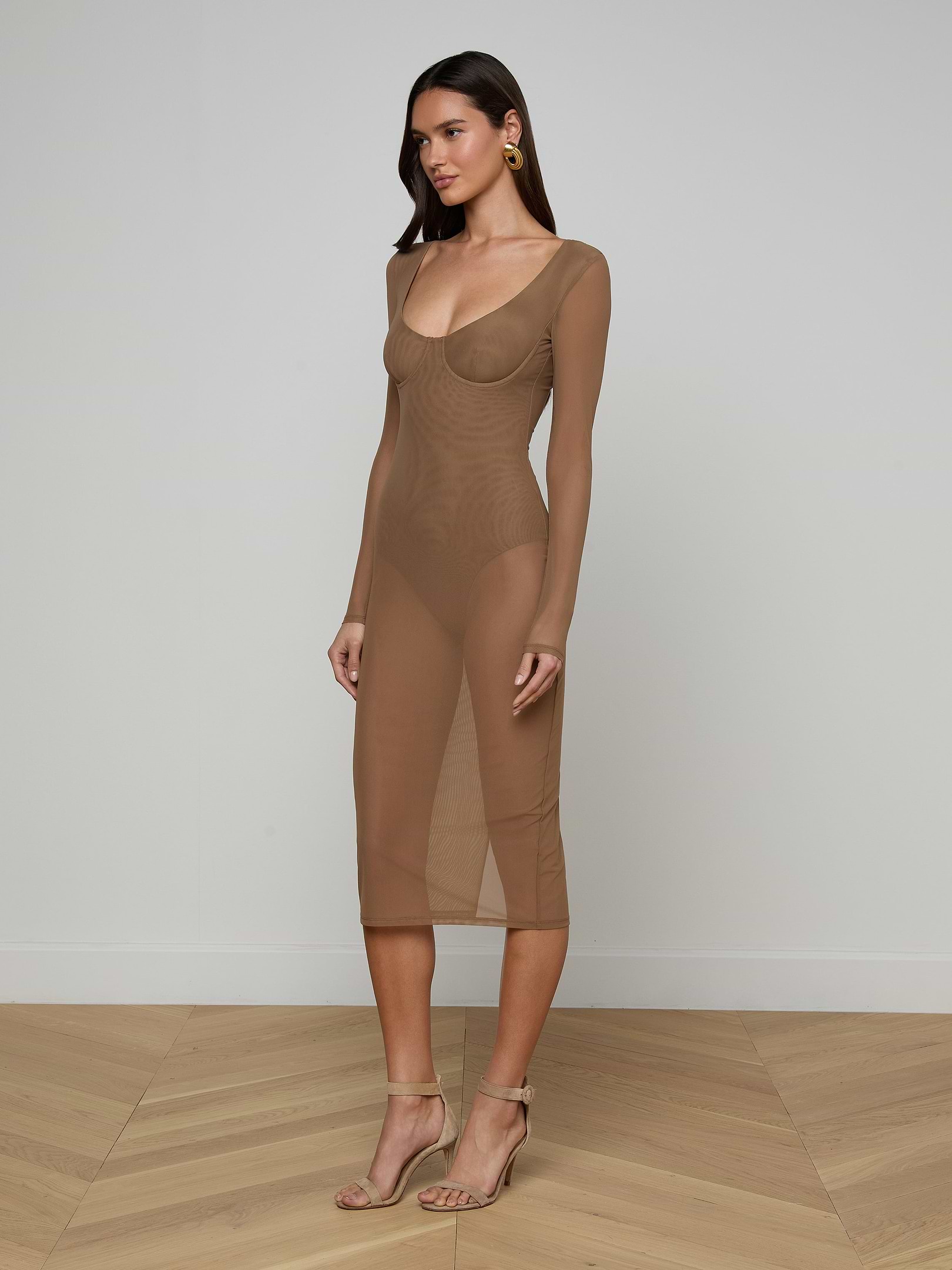 A woman stands against a plain off-white background wearing the Sorella Mesh Midi Dress—a sheer, long-sleeve, fitted brown dress crafted from stretch mesh with a bodysuit lining—paired with matching heels and gold earrings.