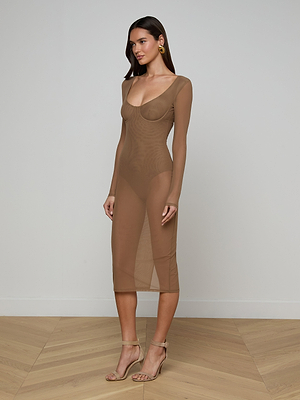 A woman with long brown hair stands in a minimalist room wearing the Sorella Mesh Midi Dress, featuring a sheer, nude design with built-in bodysuit, paired with nude heels and gold earrings.