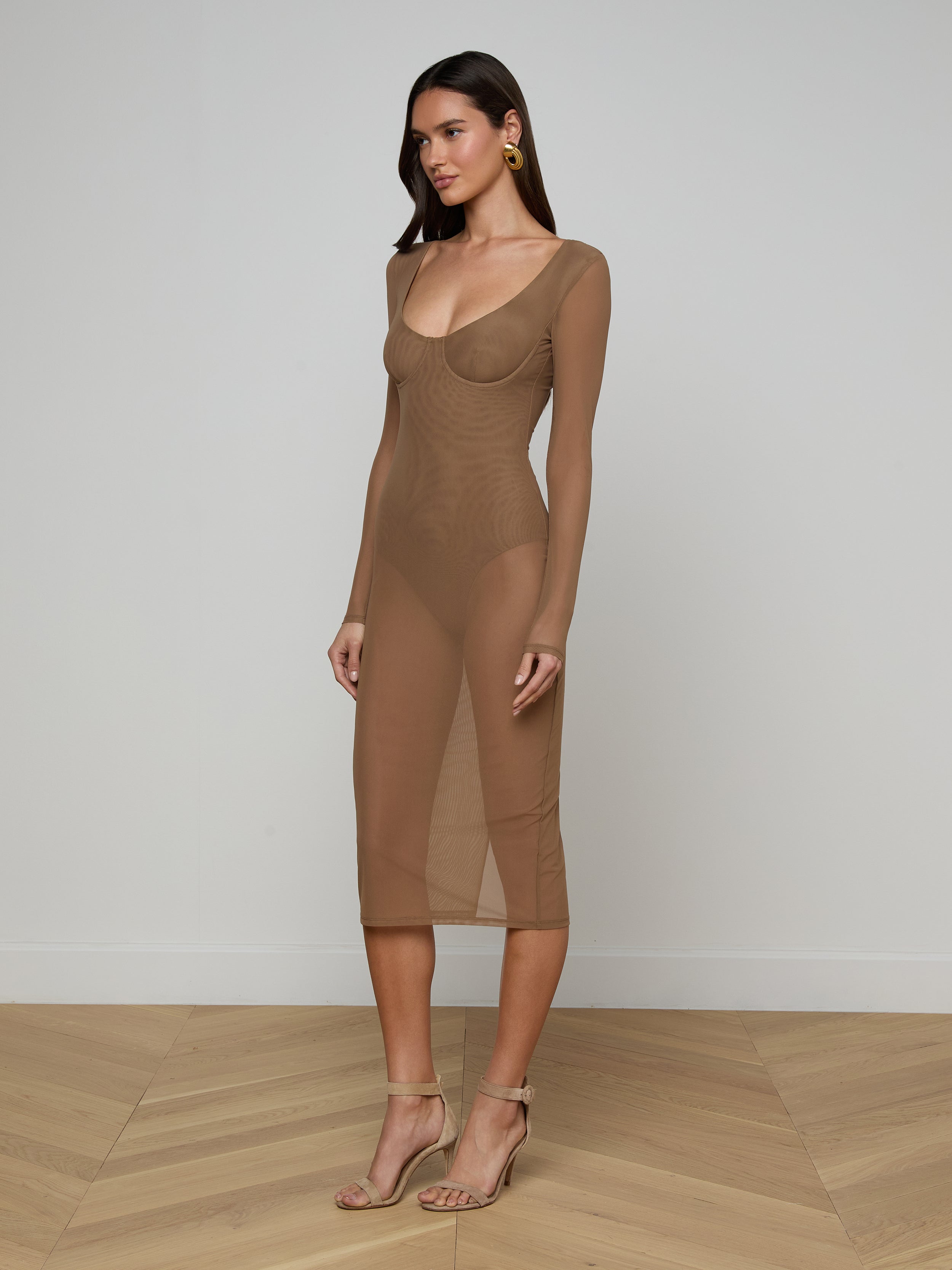A woman stands against a plain off-white background wearing the Sorella Mesh Midi Dress—a sheer, long-sleeve, fitted brown dress crafted from stretch mesh with a bodysuit lining—paired with matching heels and gold earrings.