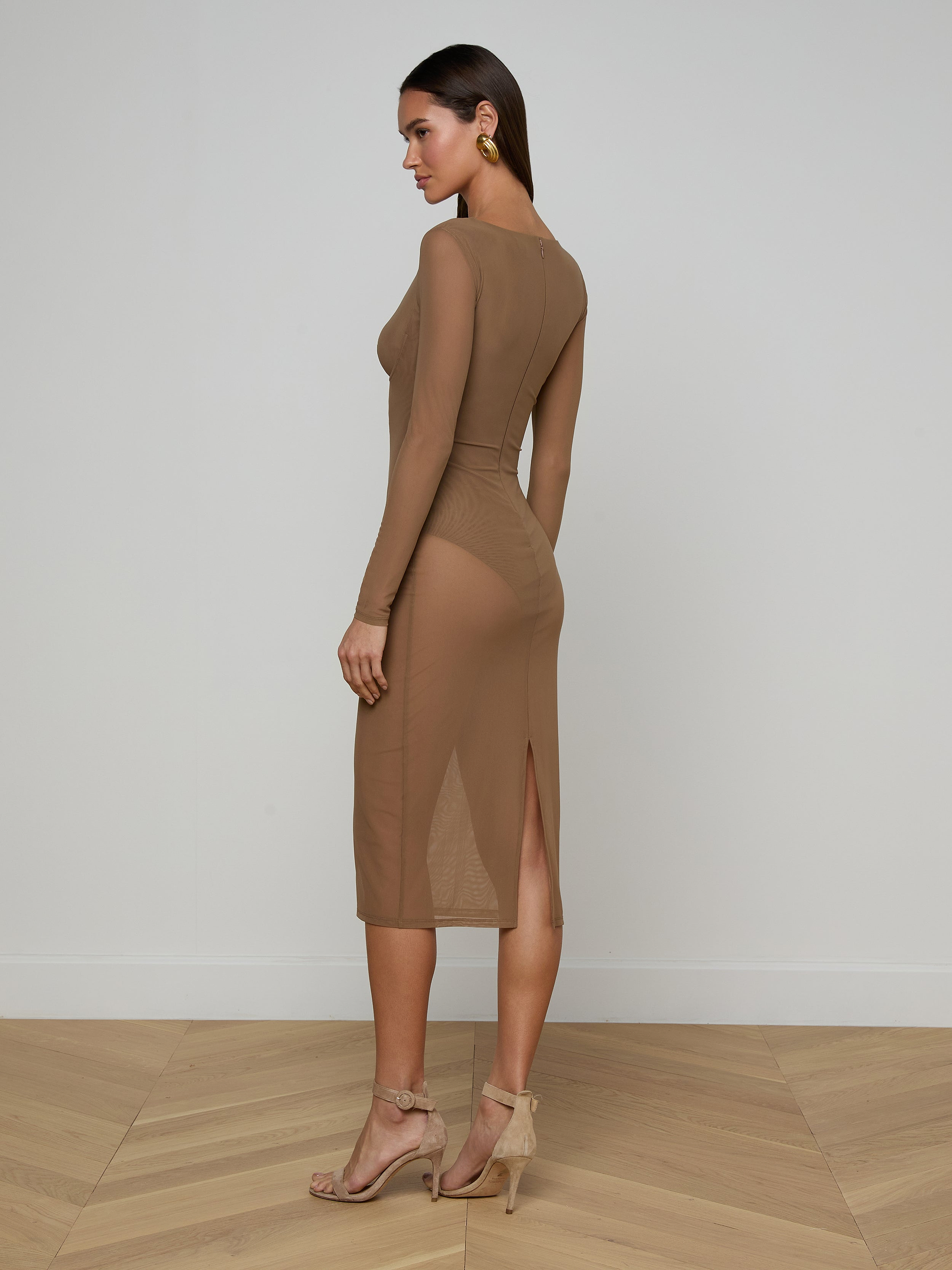 A woman poses sideways against a plain white background wearing the Sorella Mesh Midi Dress— a fitted, long-sleeve brown midi dress featuring stretch mesh and a side slit. She completes the look with high heel sandals and earrings.