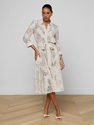 A woman wearing the Ralphie Lace Midi Dress in white—featuring long sleeves, intricate floral lace details, and a tie belt—stands against a plain light background and pairs the look with open-toed heels.