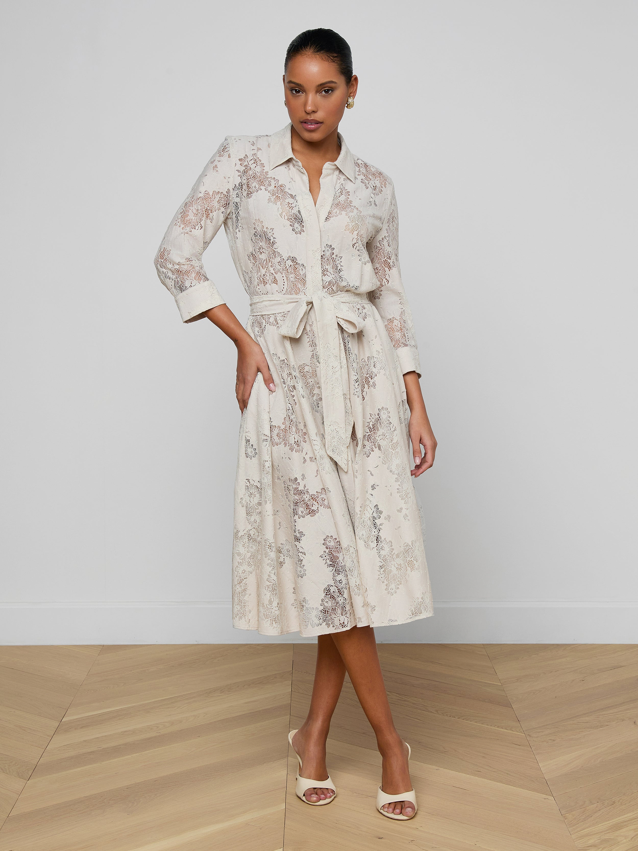 A woman wearing the Ralphie Lace Midi Dress in white—featuring long sleeves, intricate floral lace details, and a tie belt—stands against a plain light background and pairs the look with open-toed heels.
