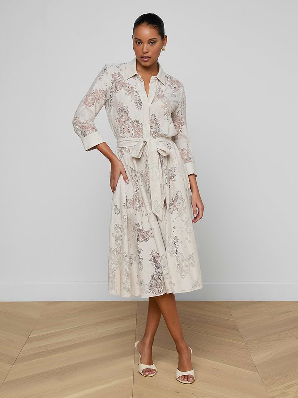 A woman models the Ralphie Lace Midi Dress, a light midi dress with a tie belt, subtle floral pattern, and rolled-up sleeves. She stands on wooden flooring against a plain white wall, wearing open-toe heels.