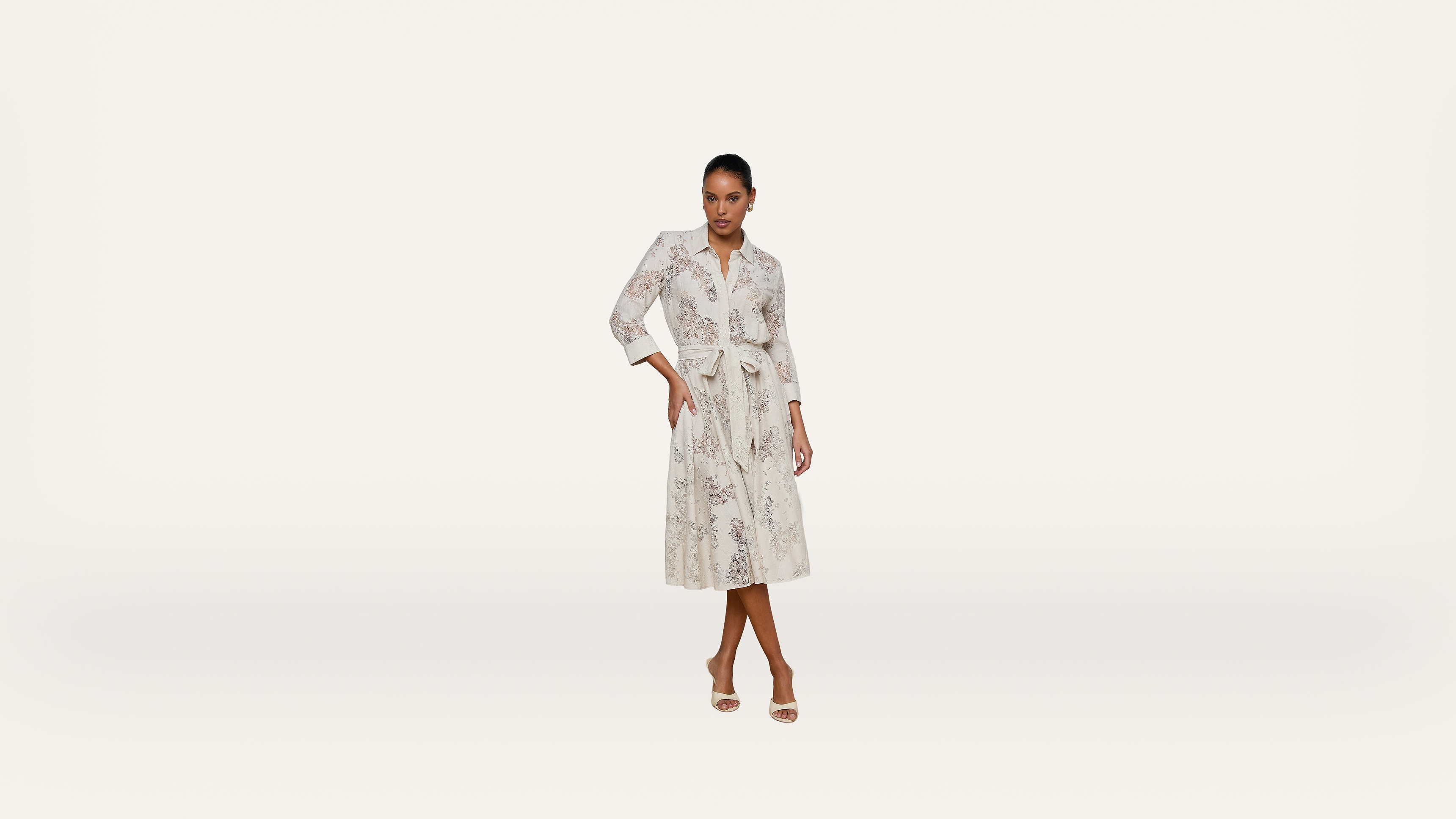 A woman stands confidently wearing a long-sleeved, belted, white midi dress with a subtle floral pattern and open-toe heels against a plain white background.