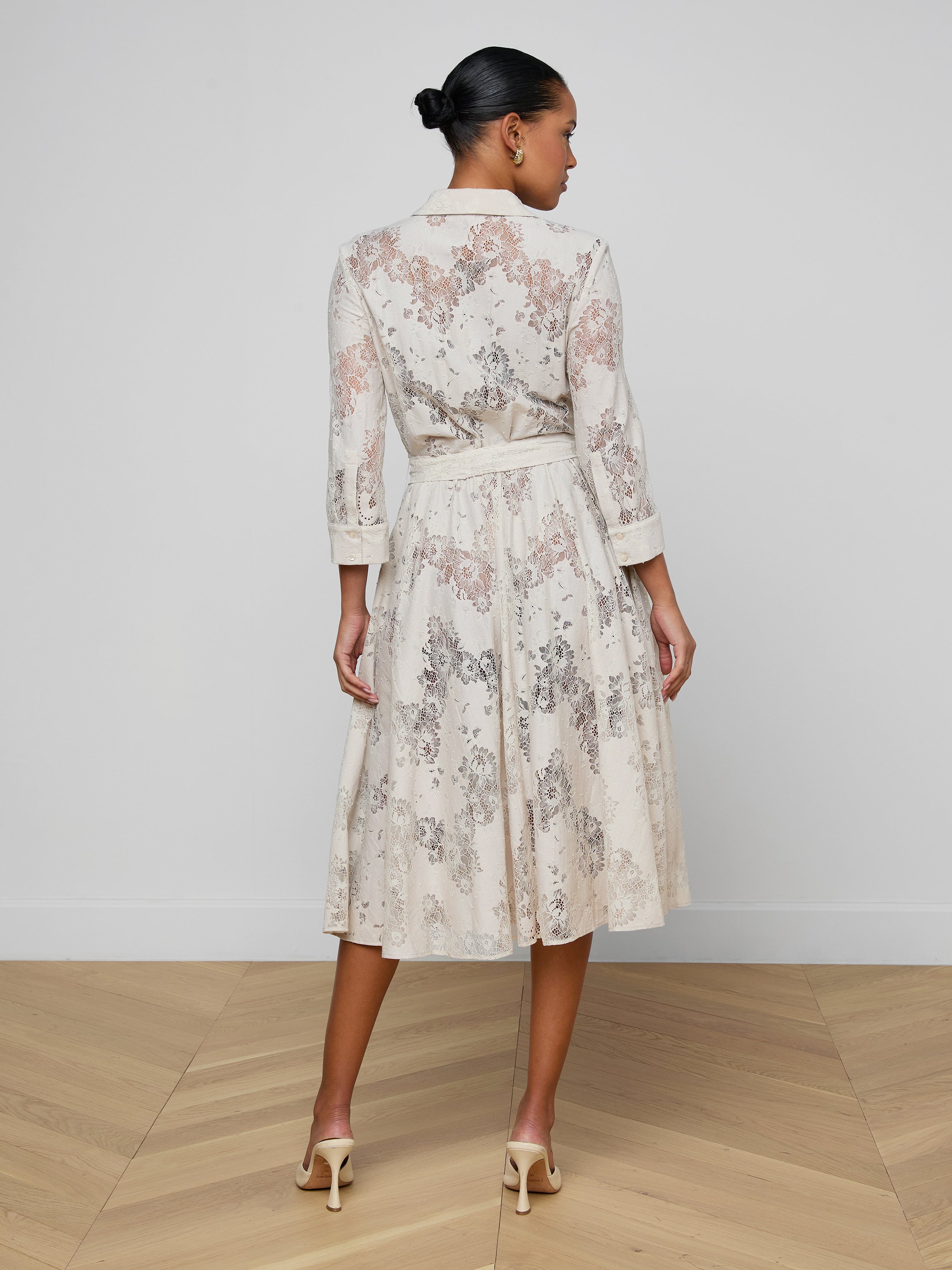 A woman stands facing away, wearing the Ralphie Lace Midi Dress—a knee-length, long-sleeved lace dress with a belt—paired with beige heels. Her hair is pulled back in a neat bun, and the background is plain white.