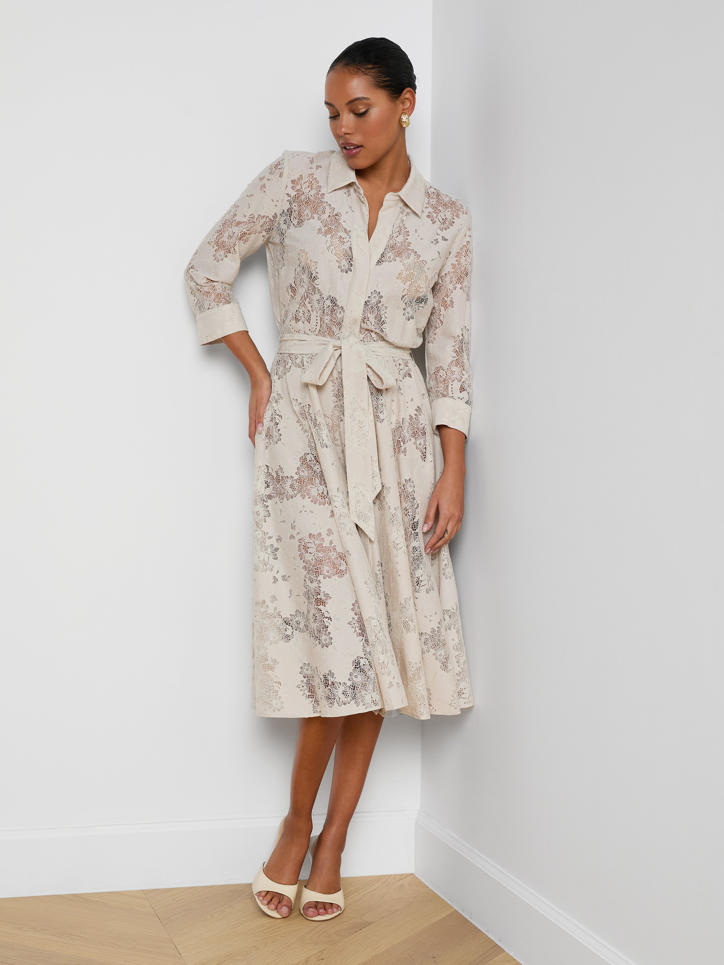 A woman wearing the Ralphie Lace Midi Dress—with a belted waist, three-quarter sleeves, and a soft floral pattern—paired with white heeled sandals, stands against a plain white background.