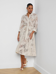 A woman wearing the Ralphie Lace Midi Dress, featuring a light beige floral pattern, long sleeves, a collar, and a waist tie, paired with nude sandals, stands against a plain white background.