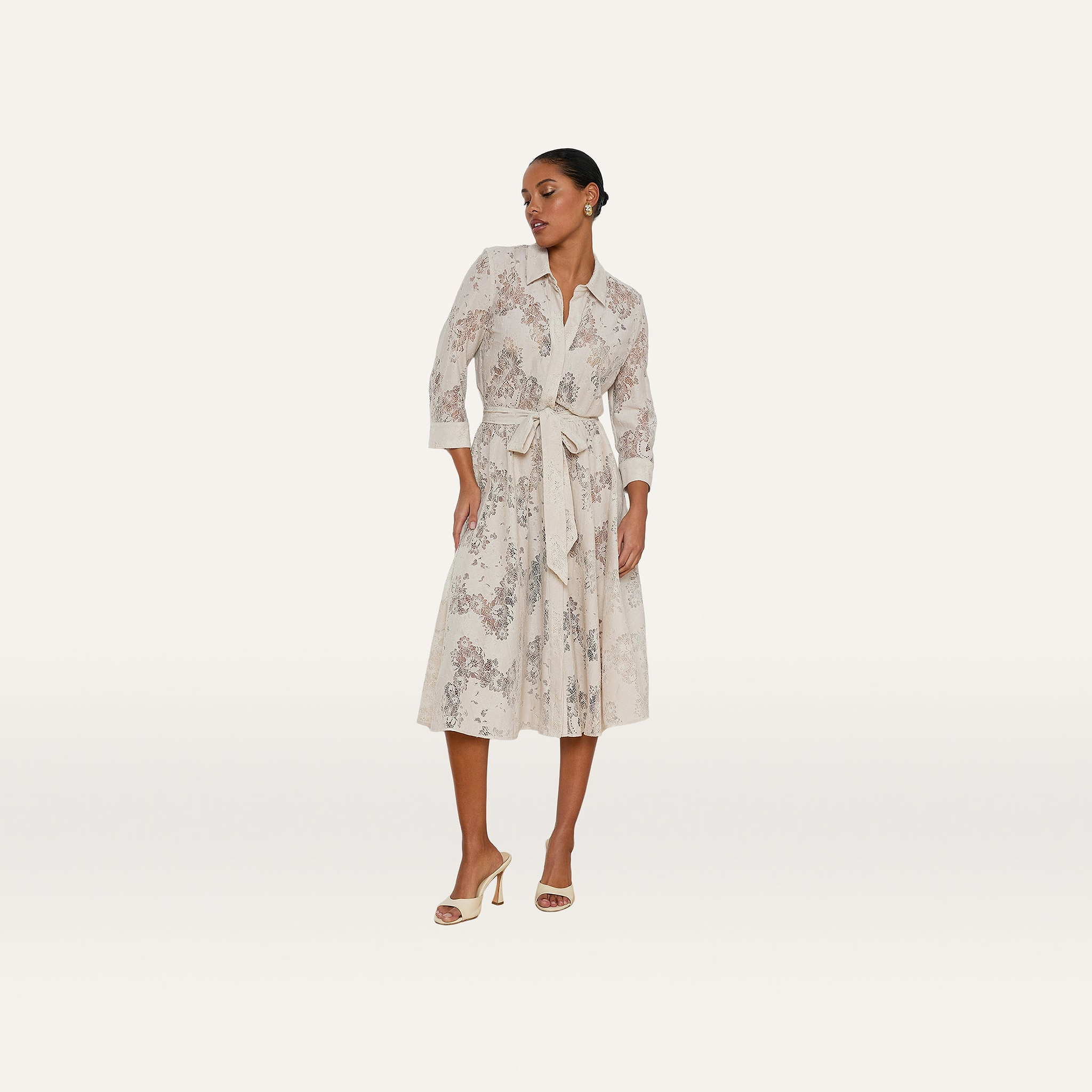 A woman wearing the Ralphie Lace Midi Dress, featuring long sleeves, a collared neckline, a tie belt, a subtle floral pattern, and a mid-length hem, paired with open-toed heels and standing against a plain white background.