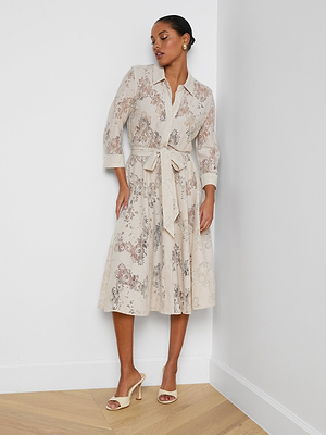 A woman stands against a white wall wearing the Ralphie Lace Midi Dress with a tie belt and beige heeled sandals. Her hair is pulled back and she completes the look with earrings.