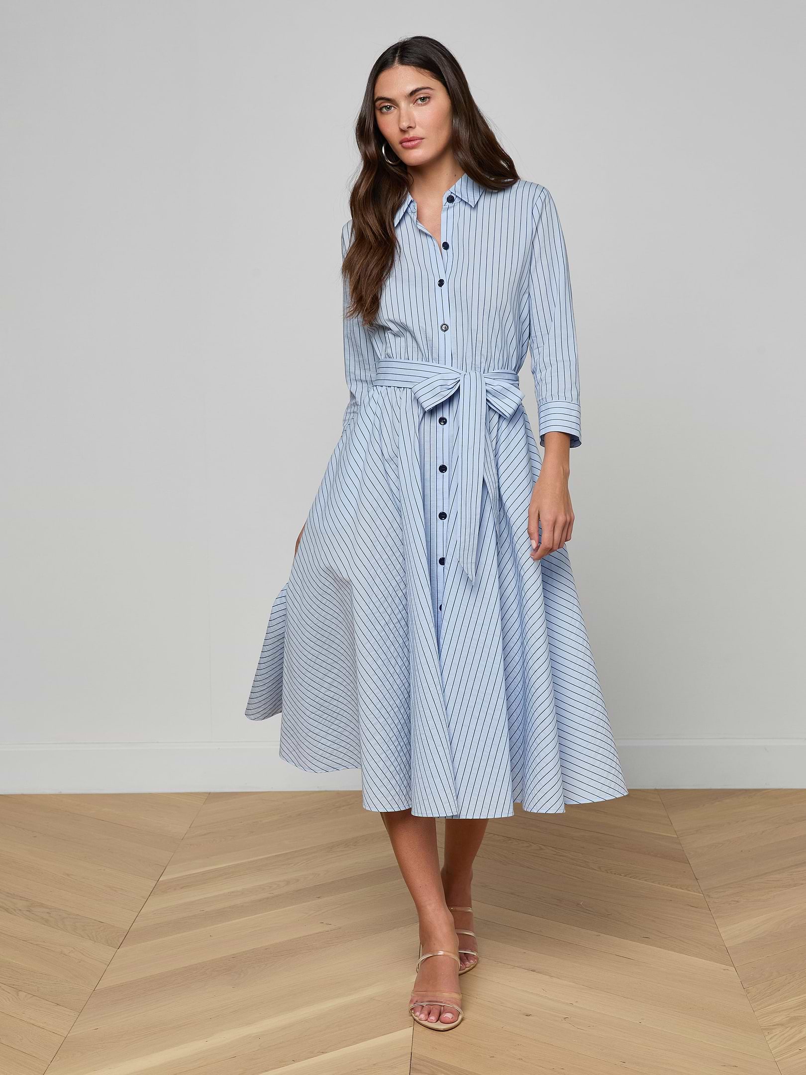 Wearing the Ralphie Midi Dress—a light blue poplin striped shirt dress with a matching waist tie and three-quarter sleeves—paired with beige heeled sandals, she stands against a plain background.
