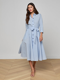 Wearing the Ralphie Midi Dress—a light blue poplin striped shirt dress with a matching waist tie and three-quarter sleeves—paired with beige heeled sandals, she stands against a plain background.