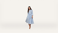 A woman stands wearing a light blue, long-sleeved, button-down dress with a tie waist and a flared skirt. She has long brown hair and is wearing sandals, posing against a plain, light background.
