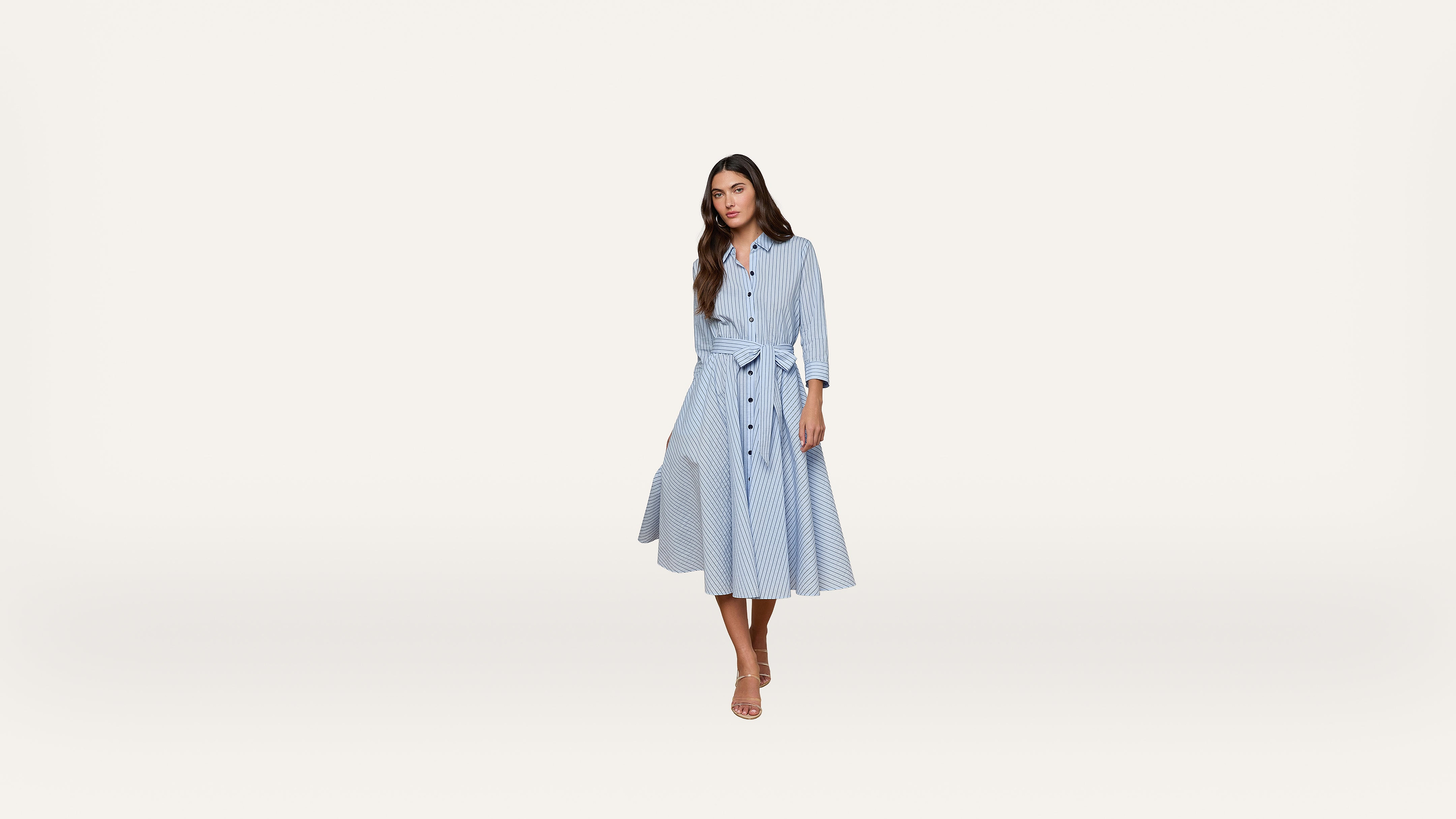 A woman stands wearing a light blue, long-sleeved, button-down dress with a tie waist and a flared skirt. She has long brown hair and is wearing sandals, posing against a plain, light background.