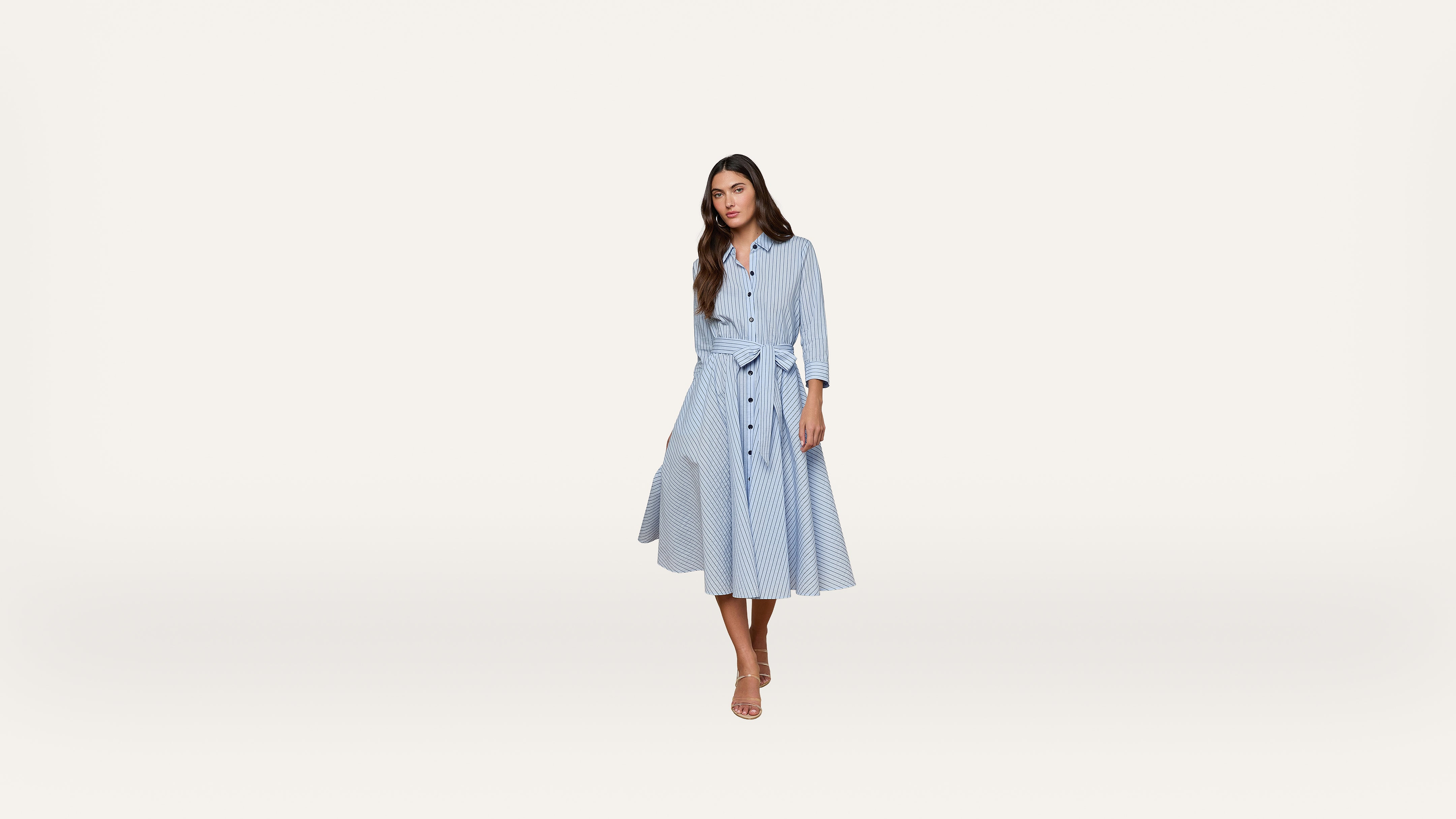 A woman stands wearing a light blue, long-sleeved, button-down dress with a tie waist and a flared skirt. She has long brown hair and is wearing sandals, posing against a plain, light background.