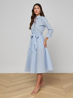 A woman stands indoors on light wooden flooring, wearing the Ralphie Midi Dress—a light blue Italian poplin striped shirt dress with a waist tie. She has long brown hair and wears clear heeled sandals. The background is plain.
