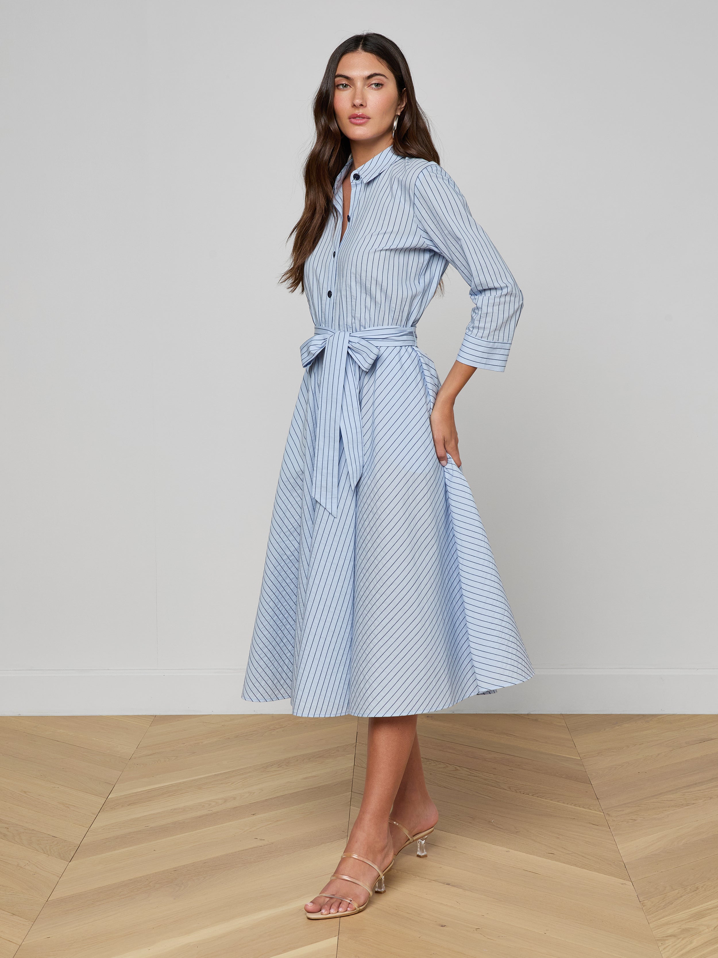 A woman stands against a plain white background wearing the Ralphie Midi Dress—a light blue poplin striped shirt dress with a waist tie—paired with nude heeled sandals.