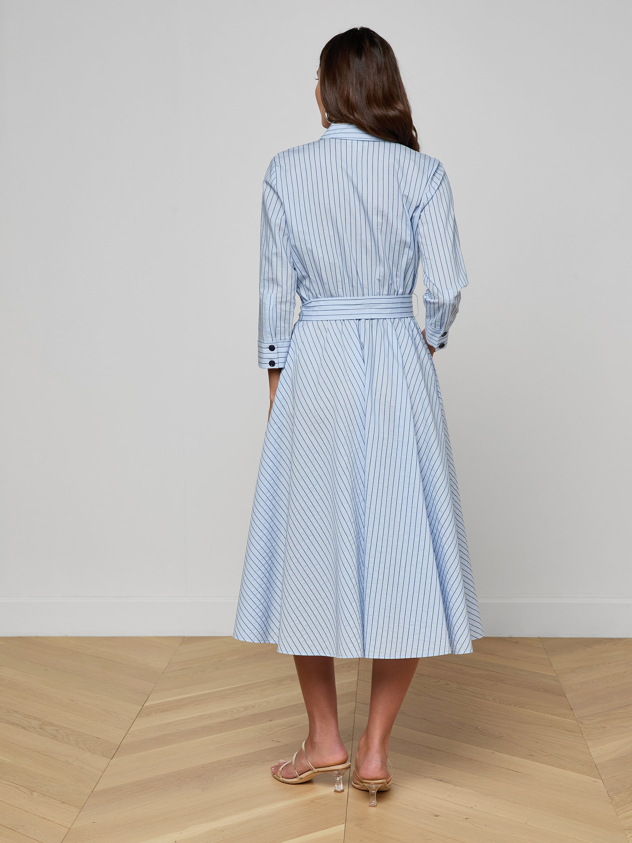 A woman stands facing away, wearing the Ralphie Midi Dress—a light blue and white striped shirt dress with long sleeves and a waist tie—paired with beige heeled sandals against a plain white background.