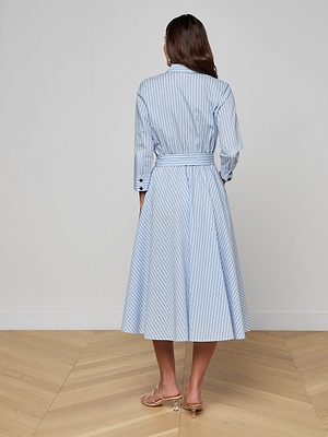 A woman stands on a wooden floor facing away, wearing the Ralphie Midi Dress—a light blue and white Italian poplin striped shirt dress with a waist tie—and tan sandals.