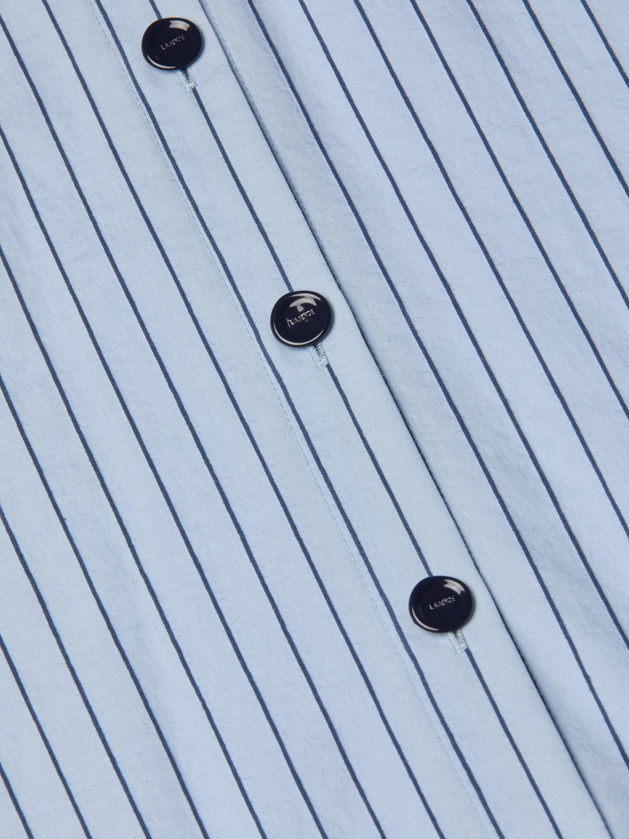 Close-up of the Ralphie Midi Dress, featuring a white fabric with vertical thin black stripes and black buttons, each labeled with LACOSTE and a small crocodile logo. The material appears crisp and neatly pressed.
